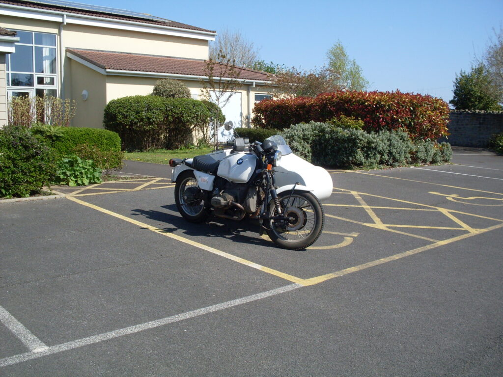 A white BMW with an open single-seater sidecar parked in the sun, in front of some bushes (with a modern low white building behind), in splendid isolation!