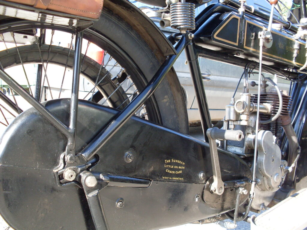A very low three-quarter rear view of a flat-tank single cylinder bike, showing part of the rear wheel, the black tank with hand change lever and linkage and very prominently the rear chain enclosure emblazoned with the words 'The Sunbeam Little Oil Bath Chain Case'