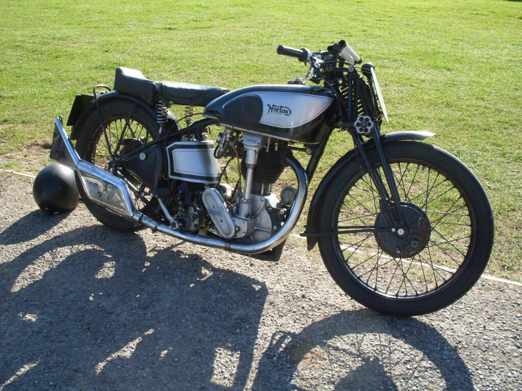 A sunny side view (against the grass) of a silver and black machine from the 1930s with girder forks and a big chrome fishtail silencer