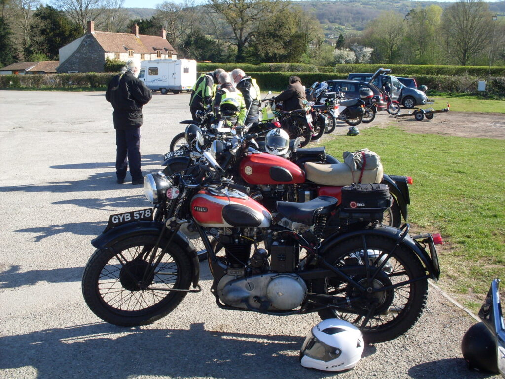 A black-framed machine with a red and silver tank in the forground with a line of machines stretching away behind, a group of riders in discussion over one of them and running across the background two hedges, trees and distant hillsides.