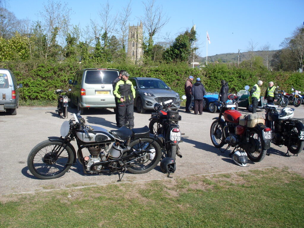 The Norton International from the other side, this time with more bikes and cars, some riders, a big hedge and a chruch tower as a backdrop.