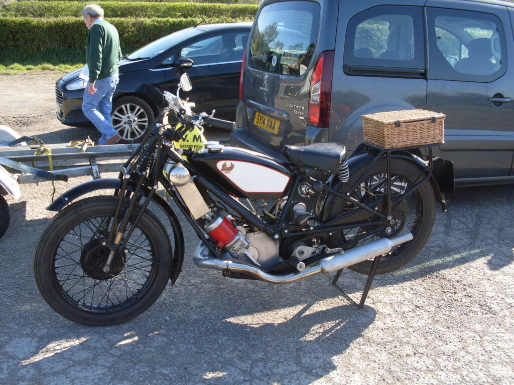 A side view of a black-framed motorcycle with a forward slanted red engine and a white-panelled back tank. It has a wicker hamper on a rear rack. Immediately behind are a trailer and two cars.