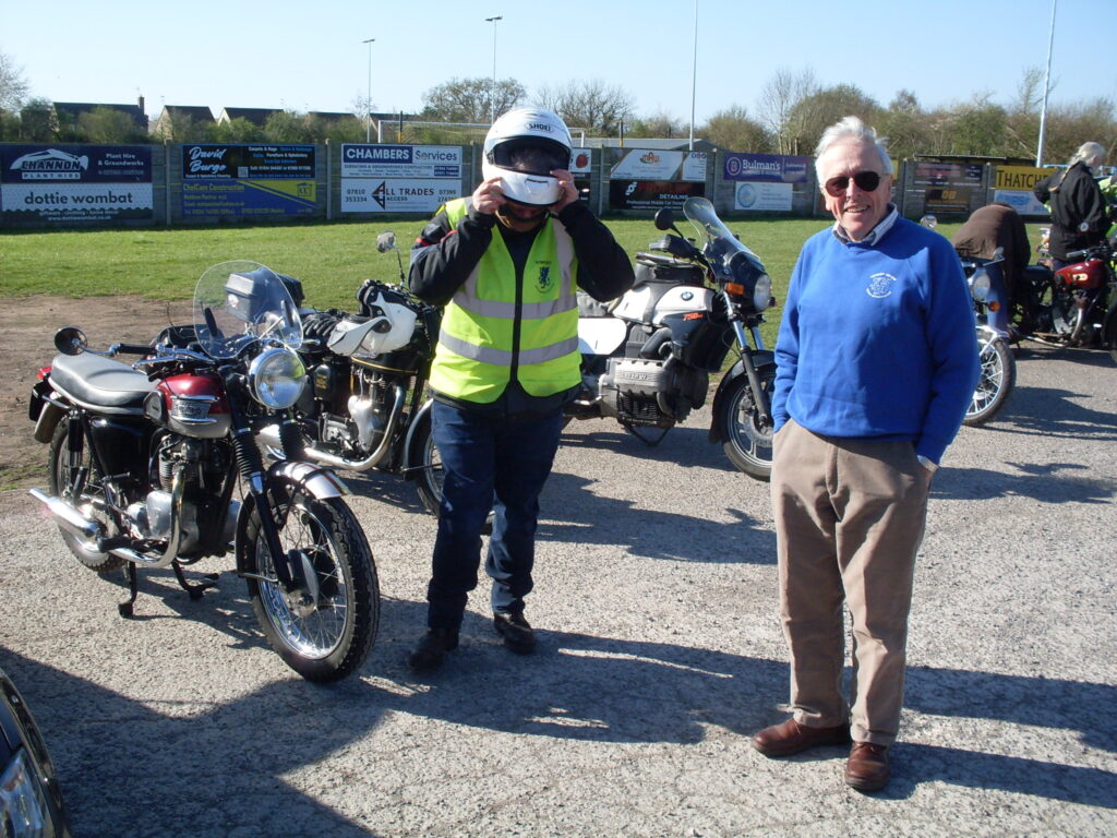 Two men standing in front of some bikes parked at the side of the ground, one in a blue top (with hands in pockets) looks at the camera, the other is in full riding gear and is just removing the helmet which is concealing his face.