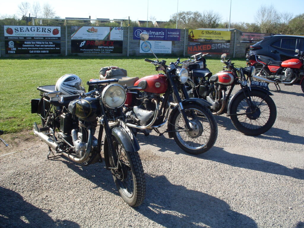 A short line-up of parked bikes, the three in the foreground are an elderly black Enfield with a big headlight, a more modern Ariel twin with a red tank and a tan dualseat and an Ariel single with a red and silver tank very much in the pre-war style of the Enfield.