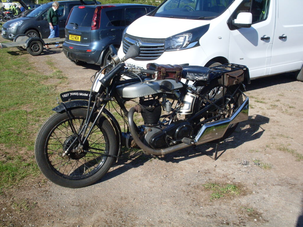 A side view of a big old Norton single, with silver-grey petrol and oil tanks, a big bulb horn on the handlebars along with a speedometer (driven from a shiny gear on the front hub) and a very shiny big chrome-plated fishtail silencer.
