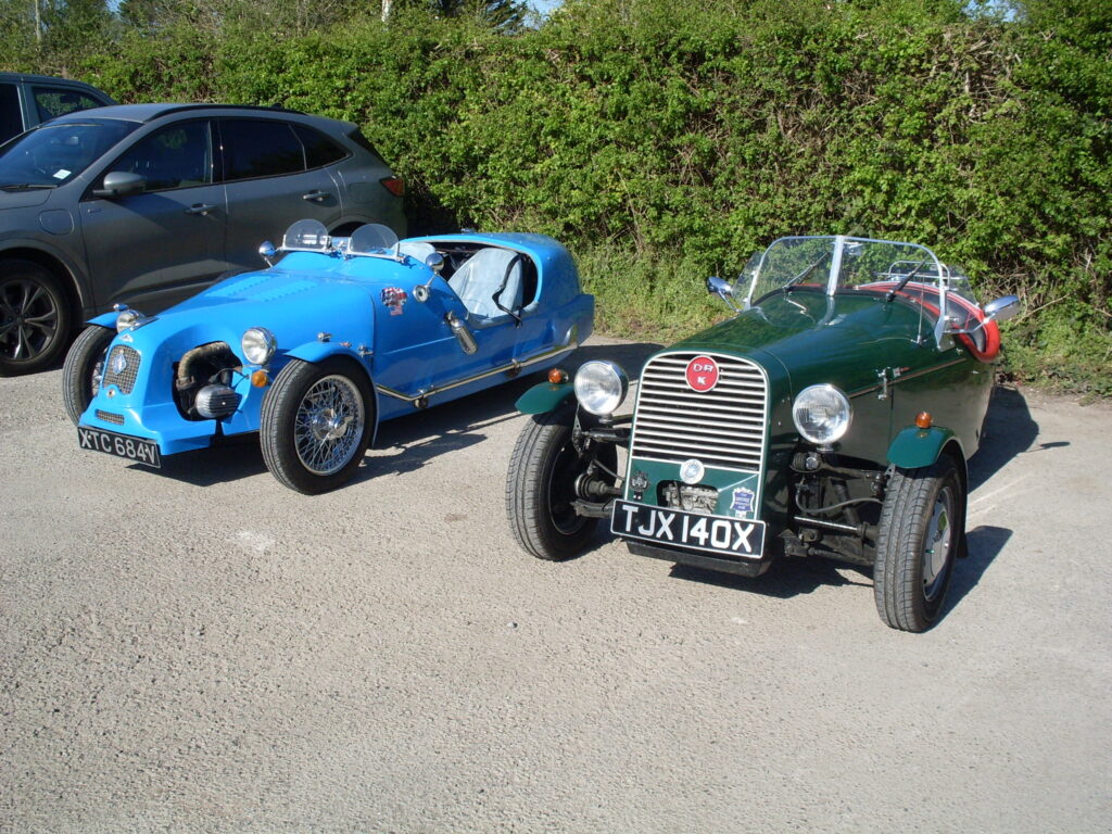 A front view of the two three-wheelers parked next to each other in front of the hedge