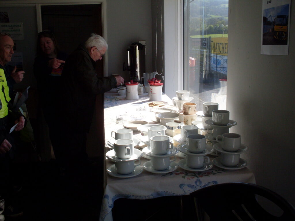Inside the Clubhouse, a long table with a 'wall' of neatly stacked clean cups and saucers in the foreground and in the background some earns and someone stirring a cup.
