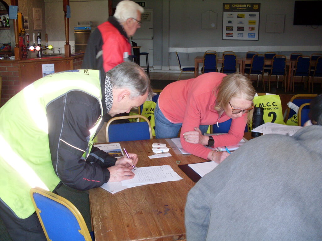 Two figures stooped over a table, on the left a man in a green bib and motorcycle gear and in the centre a lady in a red fleece, both have pens in their hands and are writing on sheets of white paper on the table.