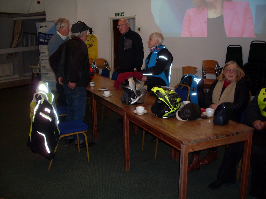 A long table with plates, cups and crash helmets on it, with people stand and sitting around it; the room seems quite dark and the camera flash has picked out the reflective strips on clothing quite dazzlingly.