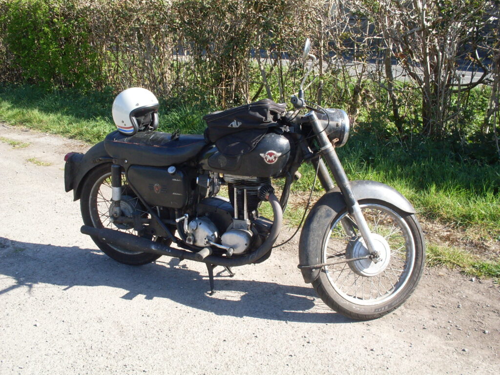 A very original and well-used looking motorcycle, with black tanks and big black mudguards. It has a black tank-bag and there is a white open-face crash helmet on the seat.
