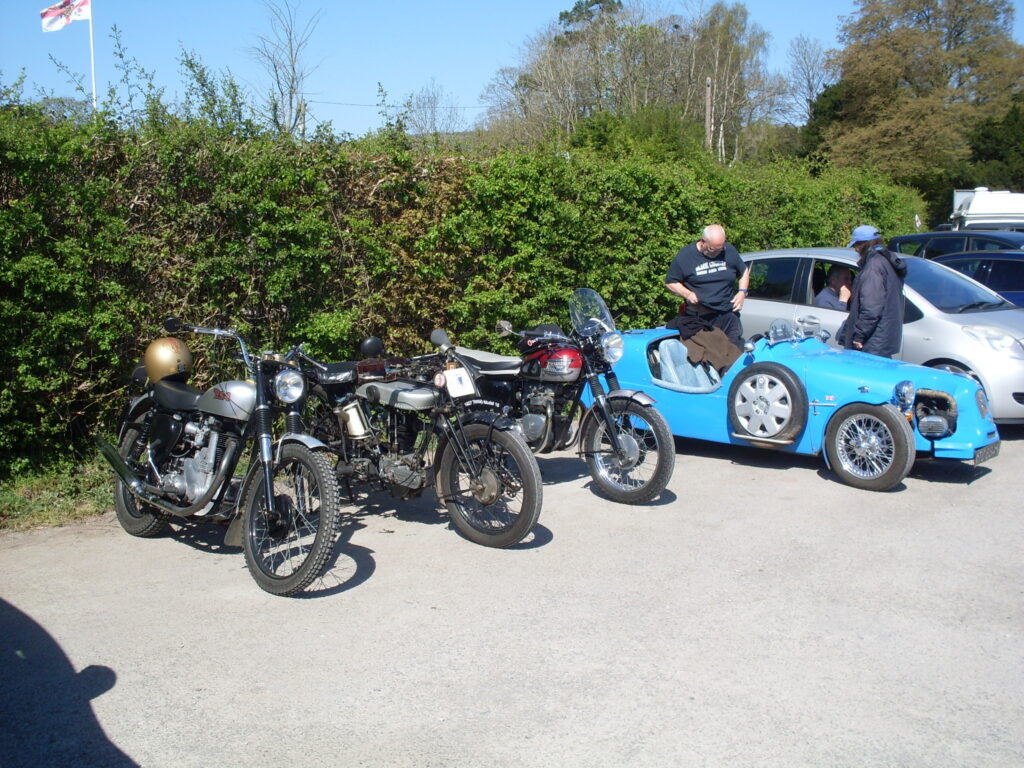 Under a bright blue sky three motorcycles and a bright blue three-wheeler are parked against the hedge. The two figures of the Lomax crew are standing behind it.