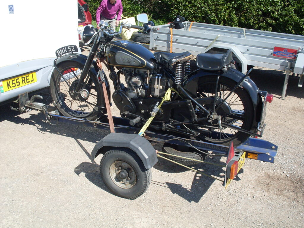 A black Velocette motorcycle strapped to a trailer behind a white van.