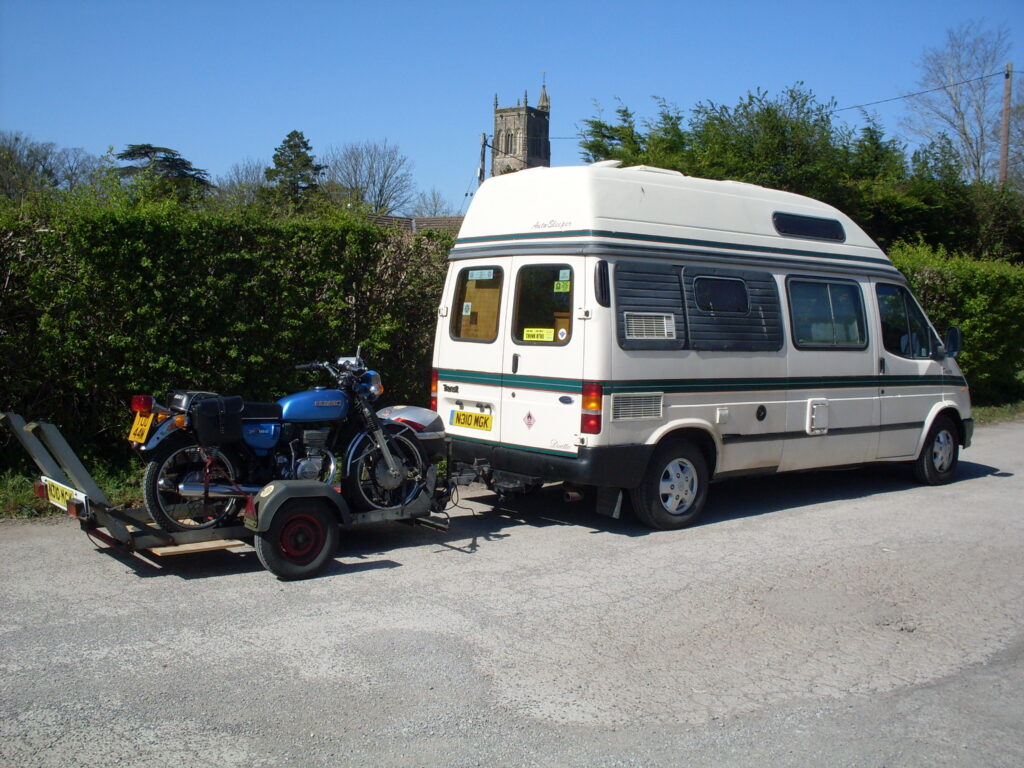 A blue Suzuki 185 from the 1970s on a trailer, behind a white campervan with green stripes.