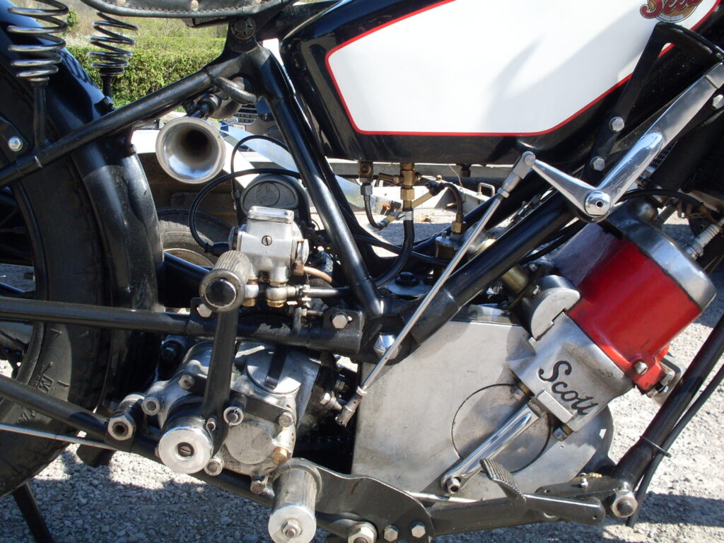 Side close-up of red Scott engine, white-panelled black tank, black frame tubes, gearbox and ancillaries with quite a few connecting pipes and unions.