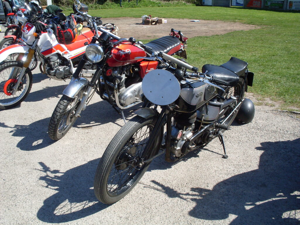 Black and silver Norton International, and red Norton Commando parked among a few other machines, in the sun.