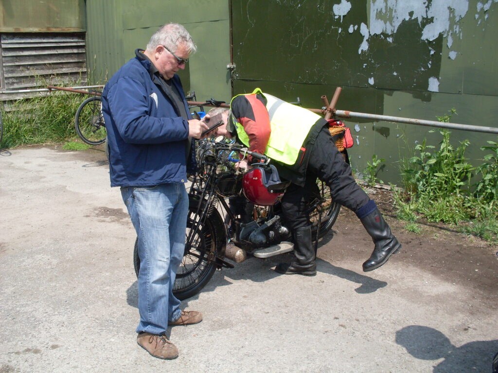 A man in blue jeans and jacket typing on a mobile phone, behind him a rider bends low over a black girder-forked machine.