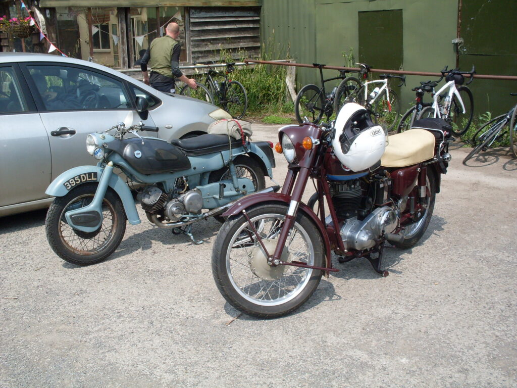 A dark red ('deep claret') single-cylinder motorbike with a beige seat parked beside a smaller grey and light blue two-cylinder machine.