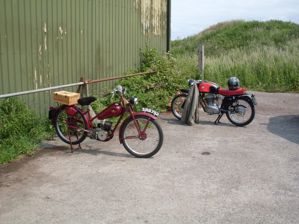Two small red motorcycles parked next to each other, the red James, with its wicker basket, parked on a rear stand nearest the camera and a very sporty and more modern  looking  machine behind it, parked on its centre stand  with a jacket gangling from the handlebars and an open-face helmet on its dualseat.