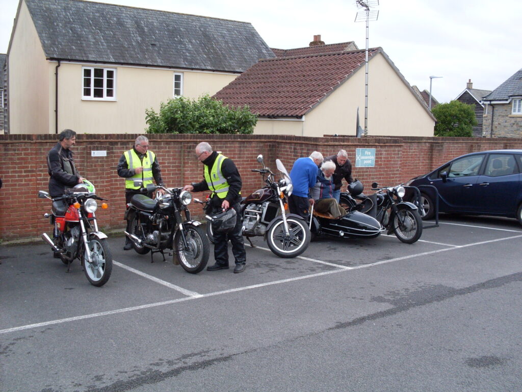 A line of several machines and riders in the car park, parked in front of a brick wall with buildings behind. at the right hand end of the line two people are helping a third figure (white haired and white bearded, with a walking stick) out of the sidecar of a black BMW outfit.
