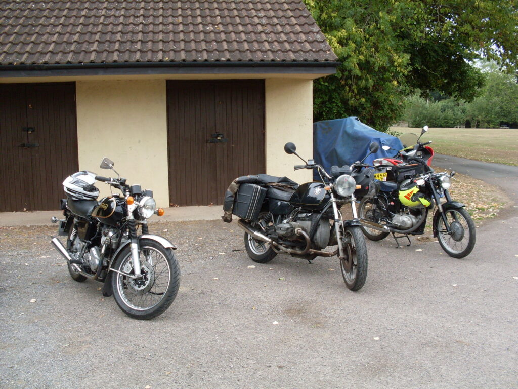 Three motorcycles parked in front of a low cream-coloured building with brown tiles and dark brown wooden double doors.