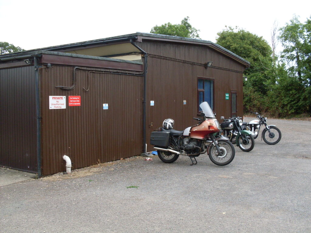 Three motorcycles parked in front of the dark brown, wooden end wall of a low single-storey building