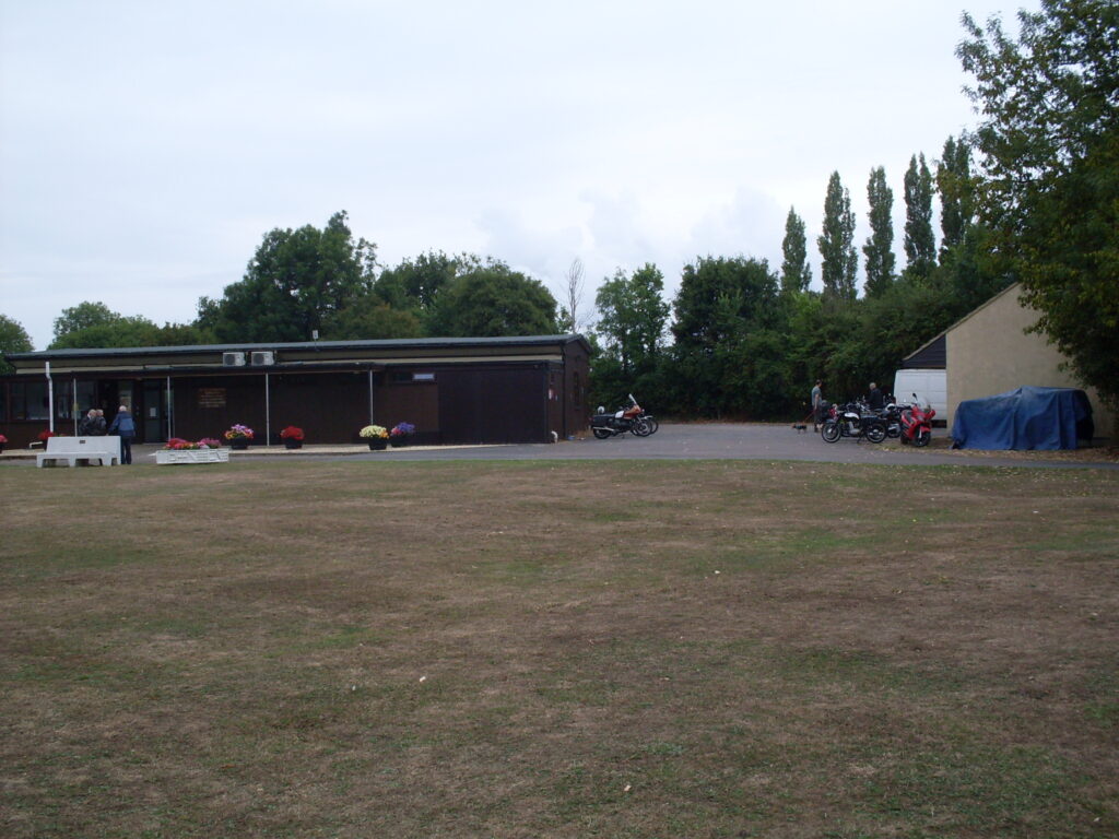 A view of a low single storey brown wooden building with the short grass of a playing field in the foreground and trees behind. There are some big planters with colourful red, yellow and pink flowers along the front of the building and a small group of figures stand talking . To the right is the cream end wall of another low single-storey building and motorcycles can be seen parked between the two. The sky is a washed-out blue with discernible clouds...