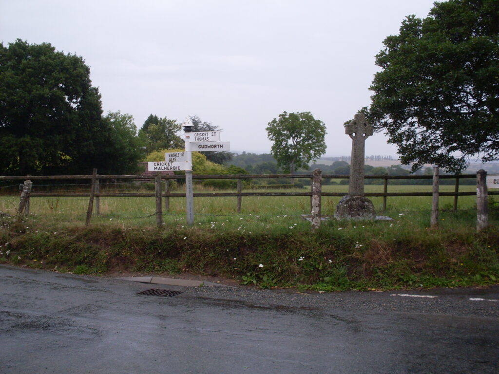 A signpost (with fingerboards) in the grassy verge at the side of a road, with trees, hedges and fields in the distance. The road surface has glistening streaks of water, the sky is grey, the horizon is misty...