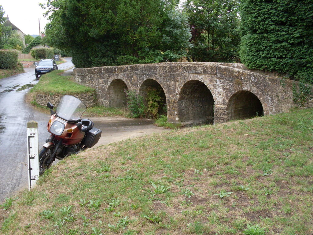 Across a high grass verge, a vertical flood-height marker board (marked up to 4 feet), a motorcycle parked immediately behind it on hard-standing that leads into the arches below a low stone bridge (obviously the alternative crossing when the water is up to the 4 feet mark!). The adjacent roadway and the hard standing are wet but with no sign of any running water.