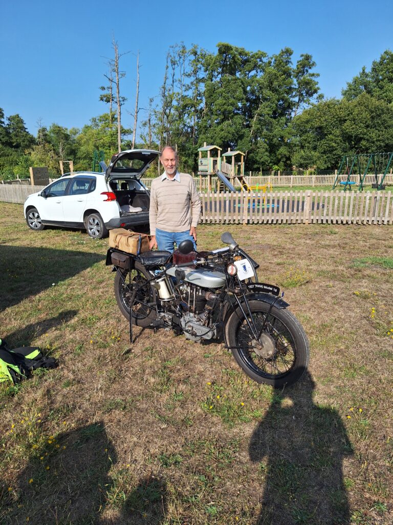 An old silver and black motorcycle stands on parched brown grass with a man in a light top and blue jeans standing immediately behind it. Behind them, a white car with the tailgate raised, a wooden picket fence, tall trees and a bright blue sky. In the foreground there are the shadows of several photographers!