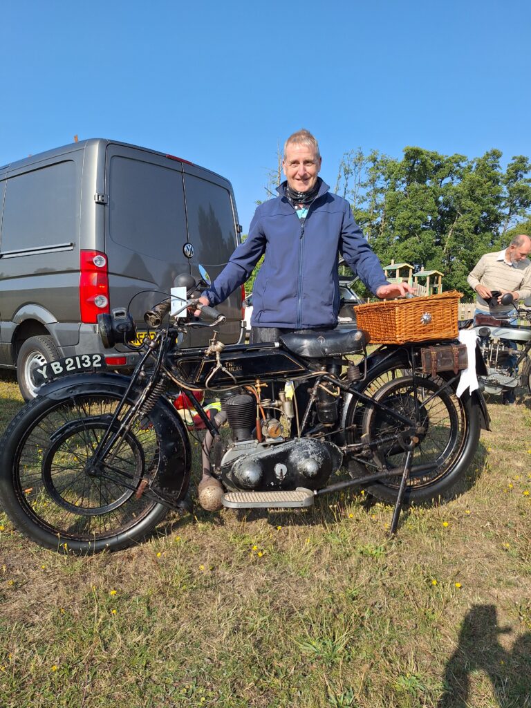 A smiling man in a dark blue fleece stands behind an old black motorcycle with a brown wicker basket behind its single saddle. Immediately behind, to one side, the back of a dark grey van looms. In the background tall leafy trees and a bright blue sky.