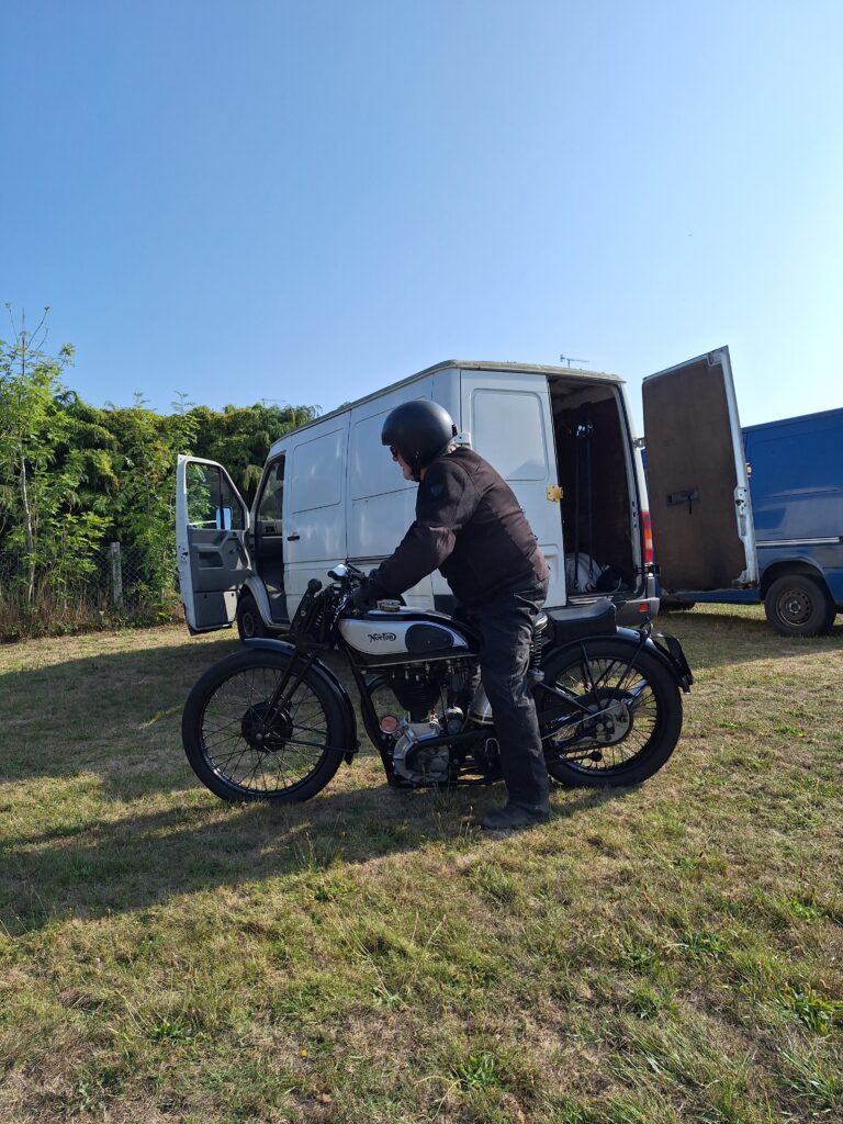 Side on, on short dry brown and green grass, a dark figure with a dark crash helmet is astride an old black motorbike with a black and silver tank. Immediately behind, rear on, a big white van with the passenger door and one back door wide open. In the background leafy trees and bright blue sky. The motorcycle and the van are casting strong dark shadows on the grass.