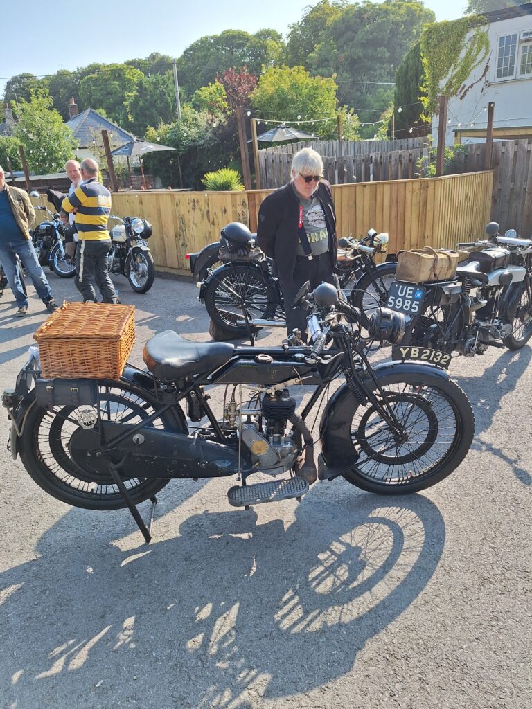 Several old motorcycles, some with baskets and boxes over their back mudguards, parked on grey gravel in front of a low light-brown wooden fence with leafy trees and a few white-painted houses behind. Several figures are moving between the motorcycles.