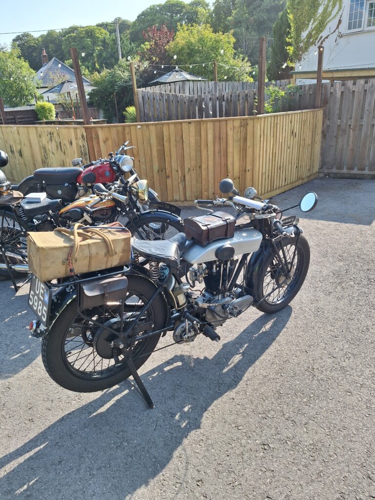 Several motorcycles parked on grey gravel in front of a brown wooden fence. Nearest the camera a black machine with a silver tank, boxy leather bags on tank and rear rack. Behind the fence can be seen a mix of houses and green trees.