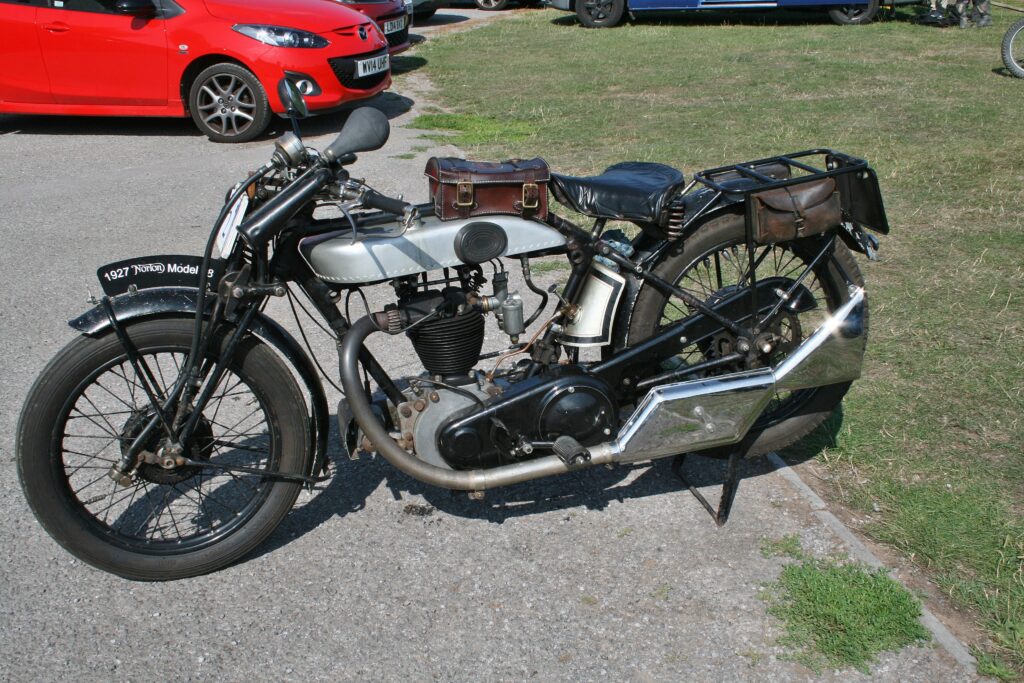 A black motorcycle with a silver tank (a small leather box strapped to it) parked on gravel, adjoining grass. The sub gleams on the big chrome-plated fishtail silencer. Behind, part of a bright red car.