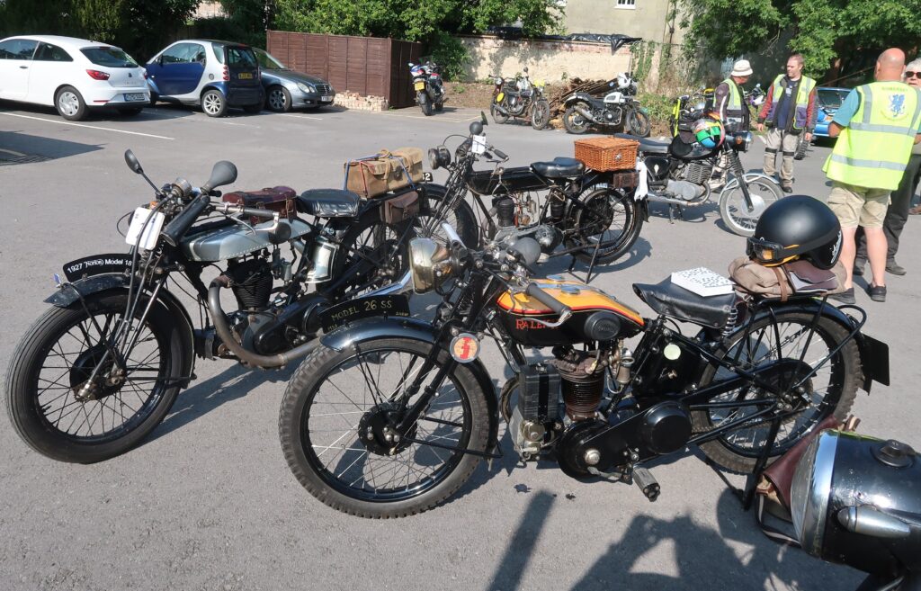Motorcycles parked on grey tarmac, with several people and some parked cars behind them. Three machines are closest, a black bike with a light tan panel to the top of the petrol tank which has 'Raleigh' in red lettering on the side, behind and to the front a black and silver Norton with a big bulb horn and leather bags strapped to its tank and rear rack , behind again a black Sunbeam motorbike with a wicker basket on its rear rack and also with a big bulb horn.