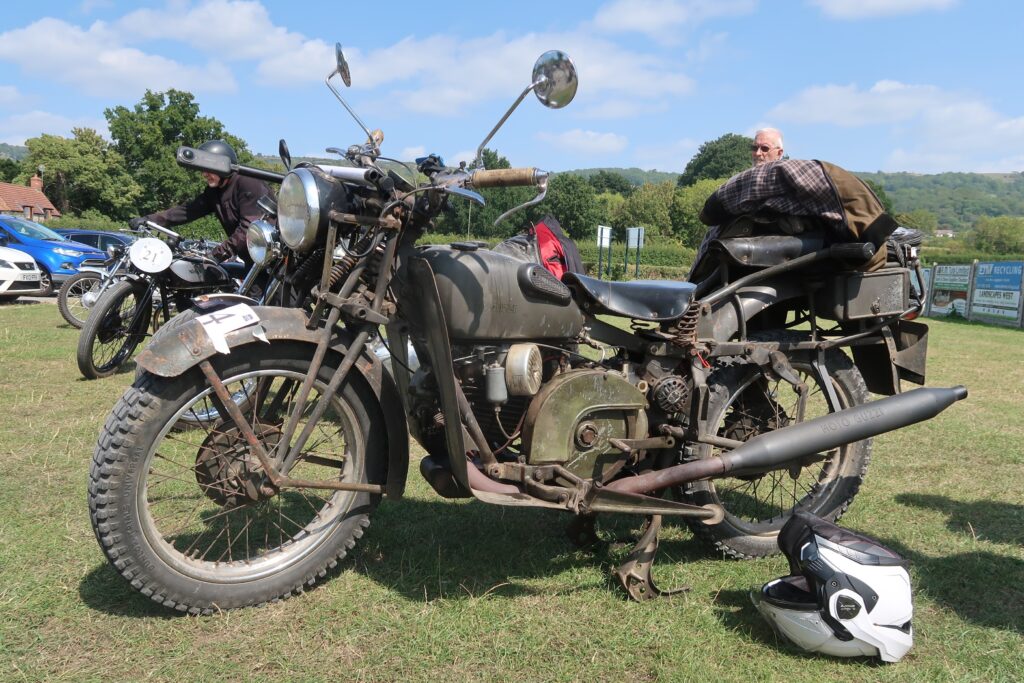 A khaki-green motorcycle with chunky tyres, legshields, single saddles for the rider and also for the pillion (the pillion seat high above the back mudguard) fills the picture. It is parked on grass, with a white helmet upside-down next to the rear wheel. There is a jacket piled on a rack behind the pillion seat, and the machine has a 'well used' look with the brown of light rust showing through the paint work. Behind are more parked cars and motorbikes, in the background trees and blue skies with white clouds.