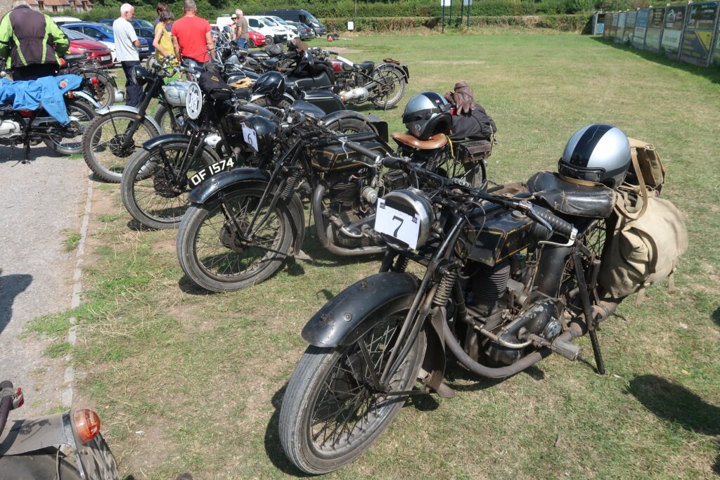 A line of black Sunbeam motorbikes parked alongside each other, on short grass. More machines, and then cars, are parked behind again. There are small groups of brightly clothed people among the bikes. The two bikes nearest the camera have canvas bags over rear luggage racks and silver and black crash helmets resting on them.