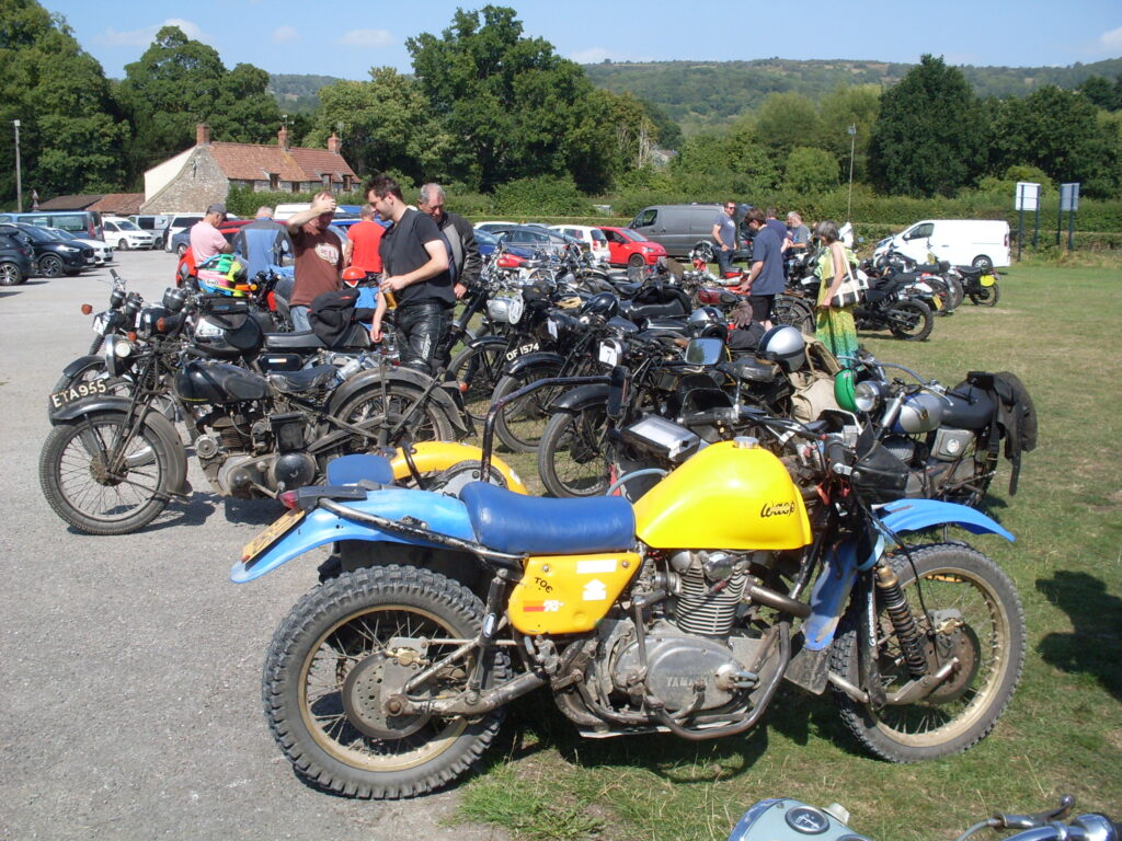 A mass of parked bikes with some parked cars behind, then in the distance a couple of red-red-tiled cottages and green hills. In front of all this a chunky sidecar outfit with blue mudguards and seat and a bright canary-yellow tank.