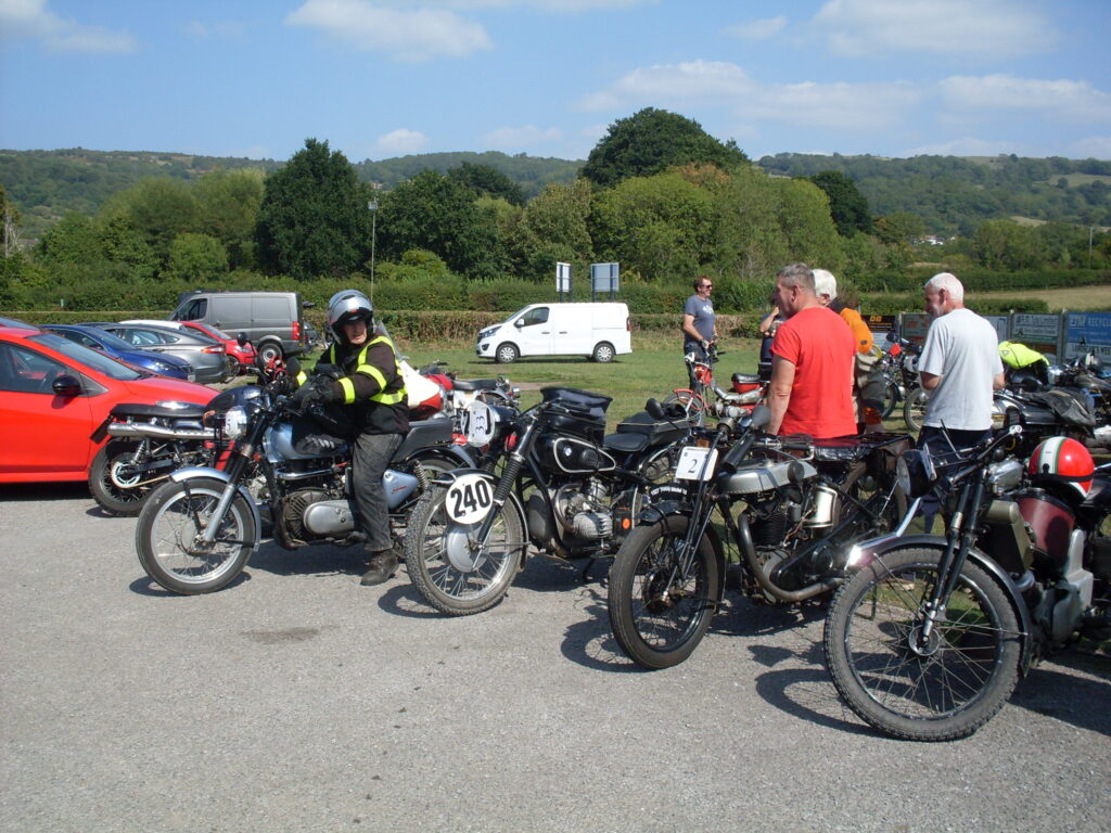 A line of 5 motorcycles, some parked cars behind. One is in the process of being ridden away, several people look on from among the parked bikes.