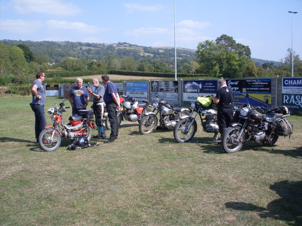 Several motorcycles parked on the grass in the sun. A small group of people are in a discussion around a small machine!