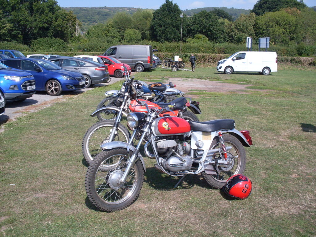 Several off-road type motorcycles parked in a line on grass, with parked cars and then trees and hills behind.
