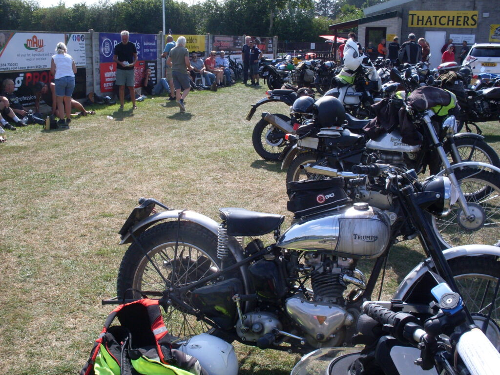 A busy scene on the grass. On one side a line of people relaxing in the sun, sitting and standing with drinks and food against a fence of advertising hoardings. In front of them, on the other side, a line of parked motorcycles , the first one that can be clearly seen a rigid-framed, alloy-engined Triumph with a chrome tank.