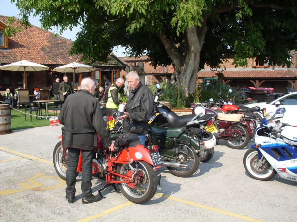 Two riders stand either side of a motorcycle, facing each other, one holding a mug of tea. Several other parked motorcycles are around them. In the background there is a big tree, several low red-tiled buildings and people sitting at tables under big sunshades.