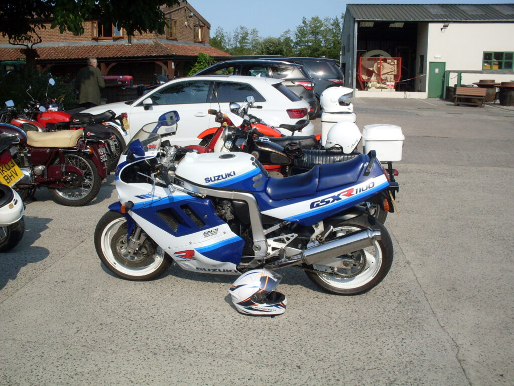 A white and blue racing-style motorcycle with white wheels, low profile tyres and big front disc brakes . Behind it several other parked motorcycles and cars.