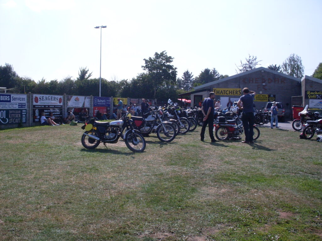 Various motorcycles parked in two lines on the grass, with people wandering among them and also sitting and standing, relaxing against a fence (with advertising hoardings). In the background a low clubhouse with a pitched roof and a prominent yellow and black 'Thatchers' sign visible on the end wall.