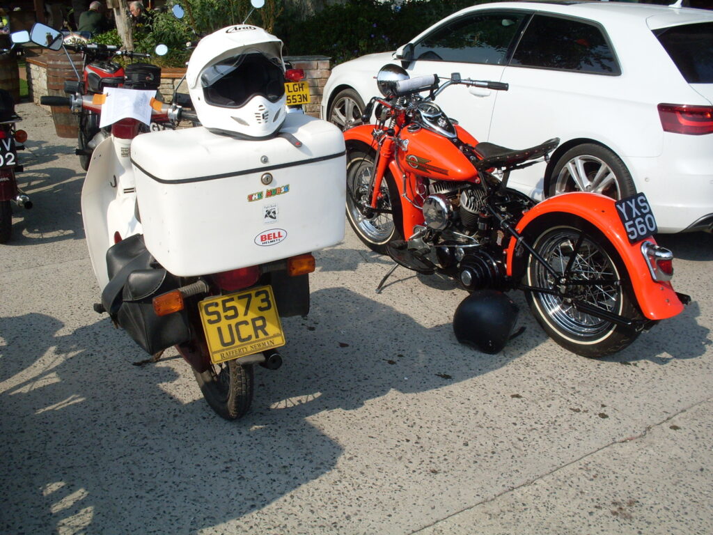 A rear view of a small 90cc Honda Cub with white legshields and an extremely large white top-box, alongside a big red V-twin Harley Davidson