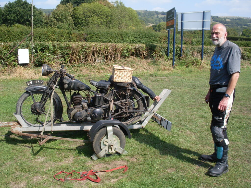 An old black single-cylinder motorcycle strapped on a trailer, on the grass. Behind the trailer a man with a grey beard, in a dark T-shirt, stands with a somewhat severe look. There is a wicker basket on the back of the motorcycle, crouched behind can be made out the shoulders and one outstretched arm of someone ion a black leather jacket.