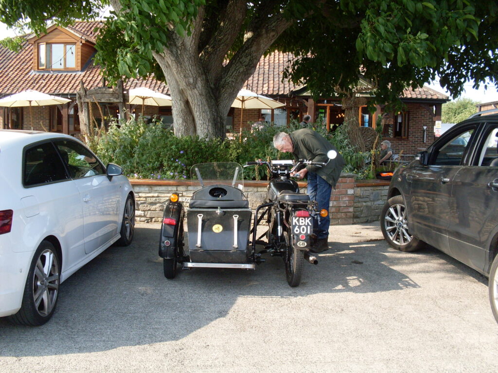 A rear view of a black BSA 650cc motorbike and sidecar, parked between two cars. Immediately behind is a low wall that is in front of some green bushes and a big tree. In the background some sunshades and a low red-tiled building. A man in a dark jacket and blue jeans is stooped low over the front of the bike.