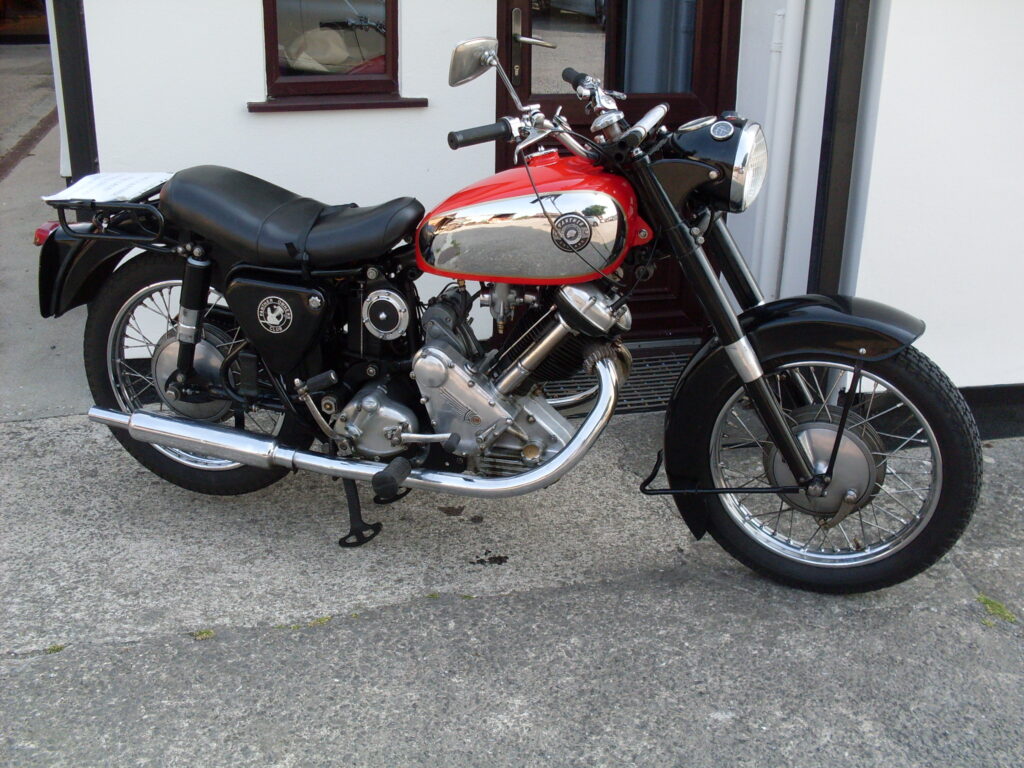 A very shiny black and chrome motorcycle, with a red and chrome tank and a tall, narrow engine sloped forward at almost 45 degrees, parked in front of a white painted wall.