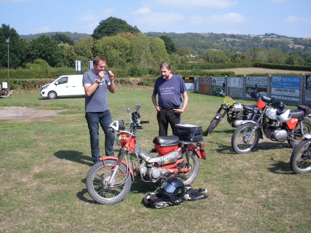 Two men in dark blue T-shirts stand behind a small red-and silver machine with a step-through frame. Around them are a couple of other parked motorcycles, behind in the distance is a white van and then trees and green hills.